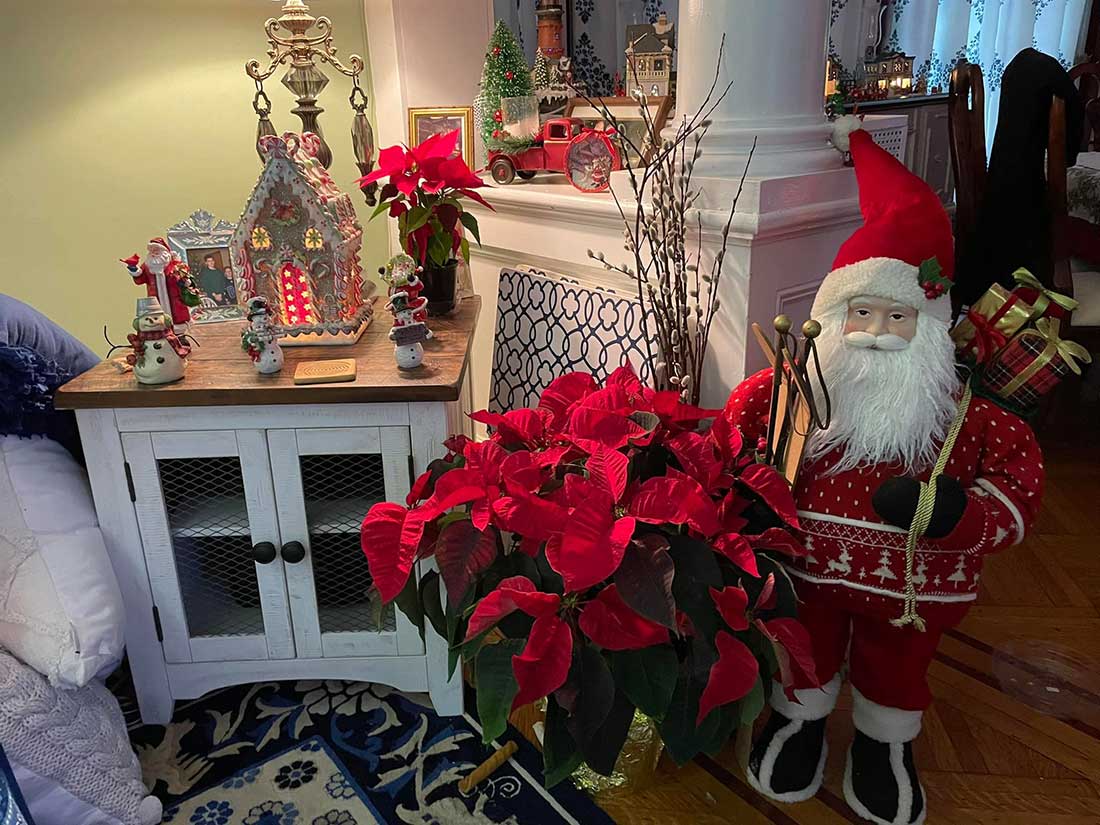 Table adorned with holiday statuary, lighted church, small poinsettia and accented with a potted poinsettia and standing Santa Claus makes for a stunning living room display.